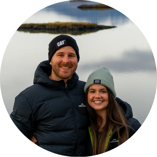 Couple standing in front of lake, islands in the background. Male and female are both wearing beanies and large jackets.