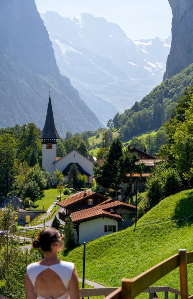 A woman in a white dress stands in the foreground overlooking a quaint village with a church and steeple, set against the backdrop of towering green mountains and valleys.