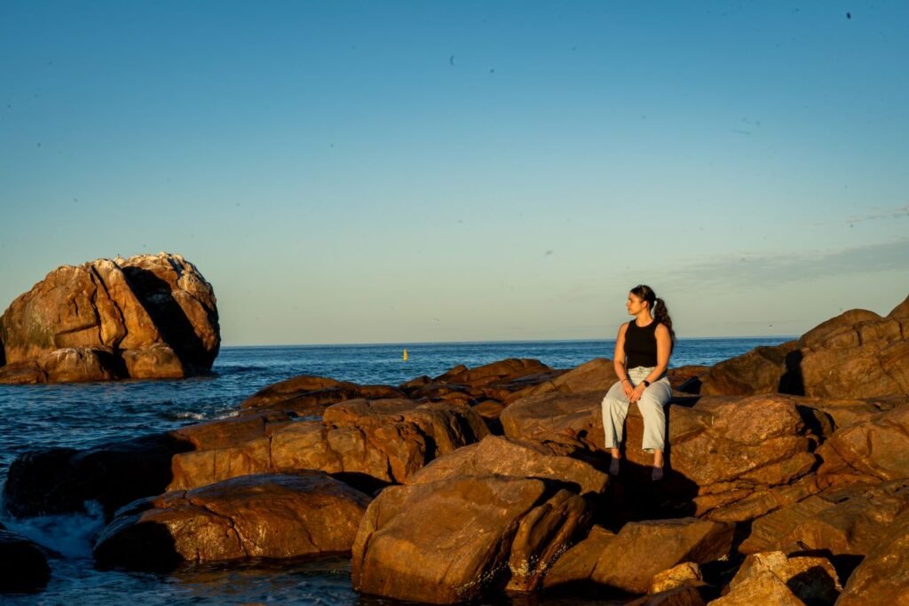 Girl sitting on rocks at sunset, staring at larger rock in distance, ocean nearby