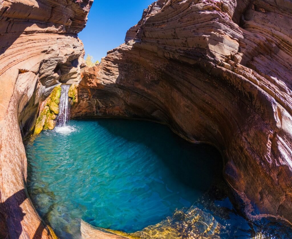 A natural rock pool with crystal-clear blue water, surrounded by layered red rock formations, with a small waterfall cascading into the pool, under a bright blue sky.