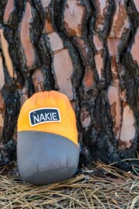Bag with orange top and black bottom that says Nakie, textured bark of tree in the background, bag sitting on straw